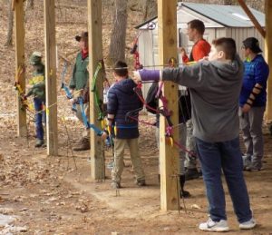 This image shows three children standing under a structure supported by wooden beams. All of the children are holding archery bows. The closet child has the string pulled back and is aiming the arrow.