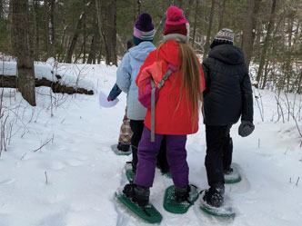 Three kids walking in a snowy woods with snowshoes on