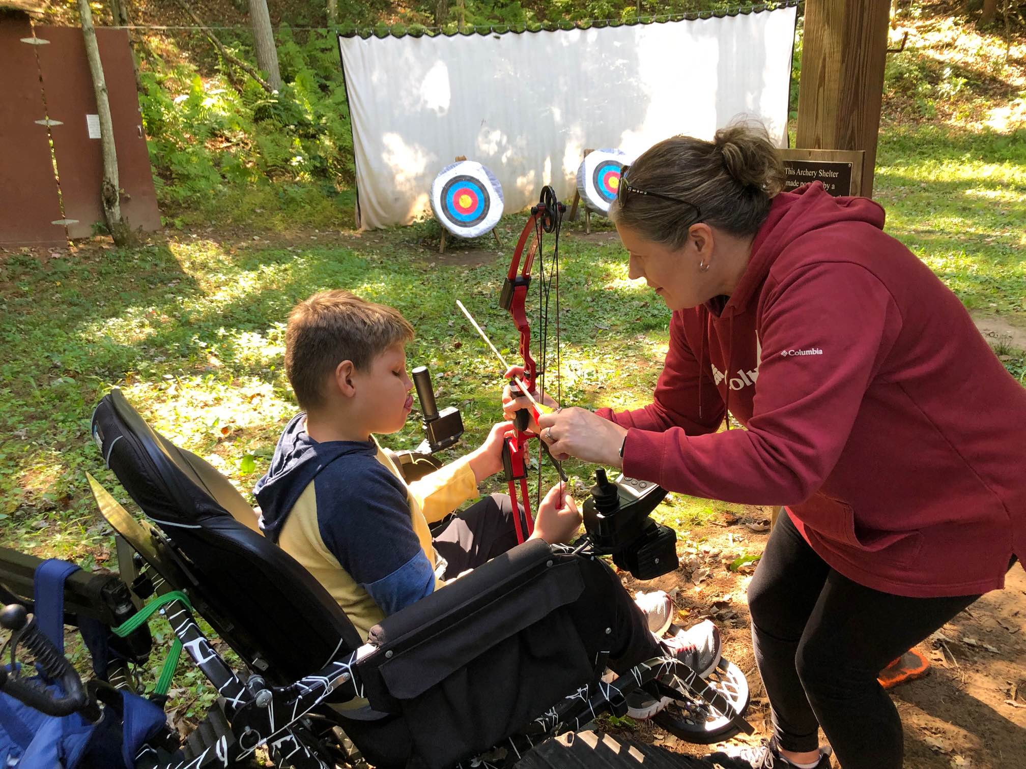 An aide helps a youth in a wheelchair shoot archery.