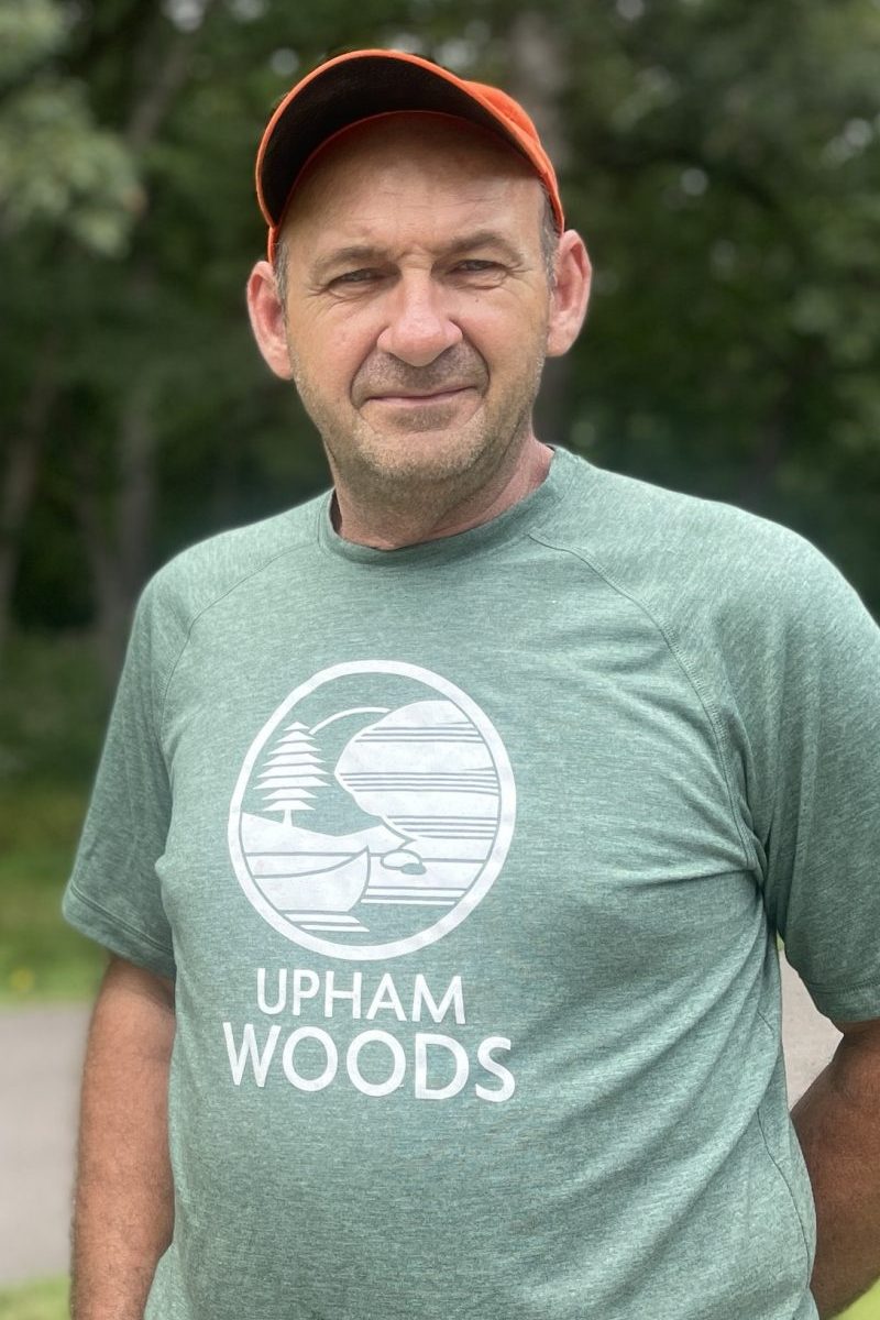 Man wearing orange baseball cap and green Upham Woods t-shirt standing next to a small tree