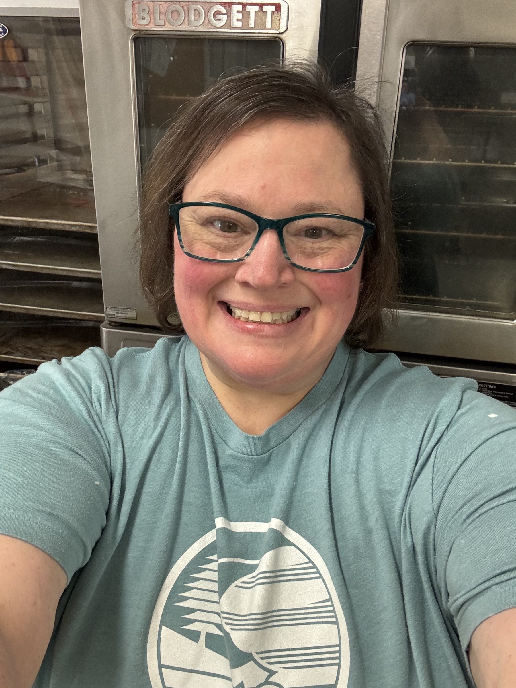 a smiling woman wearing glasses and an Upham Woods t-shirt in front of commercial ovens