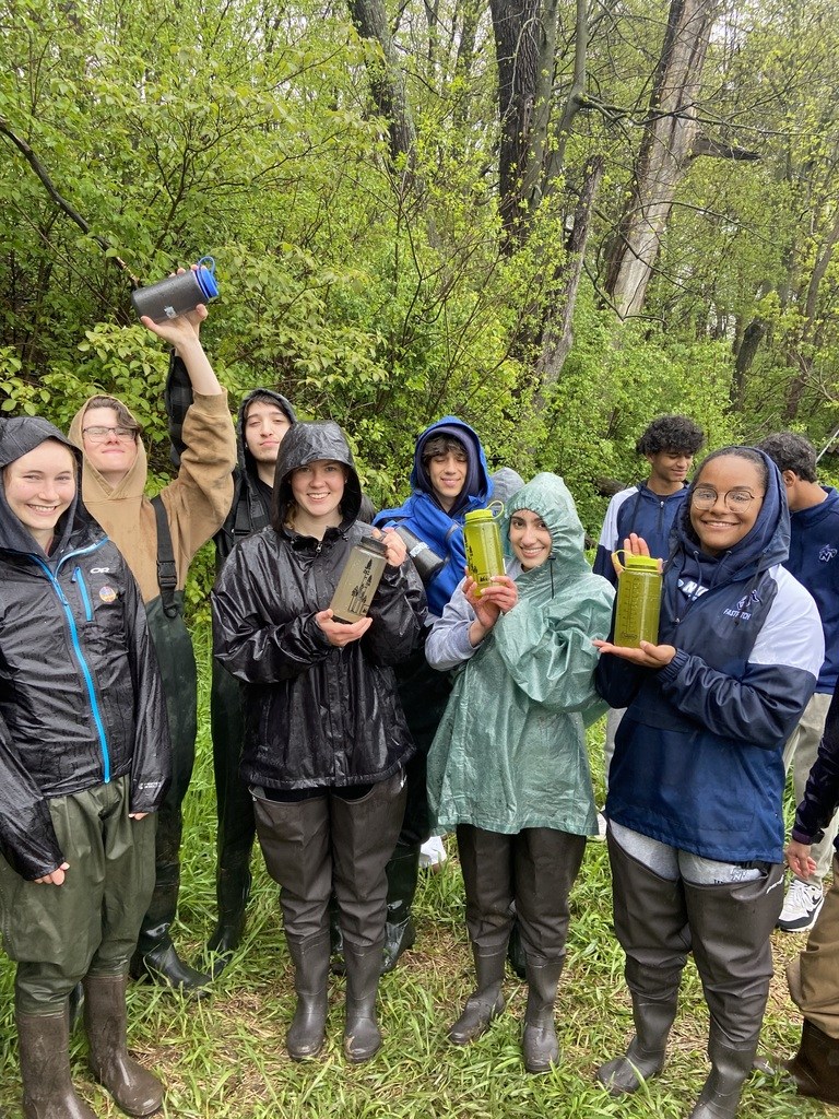 Group of smiling students wearing rain gear hold up Nalgene bottles containing Milwaukee river samples